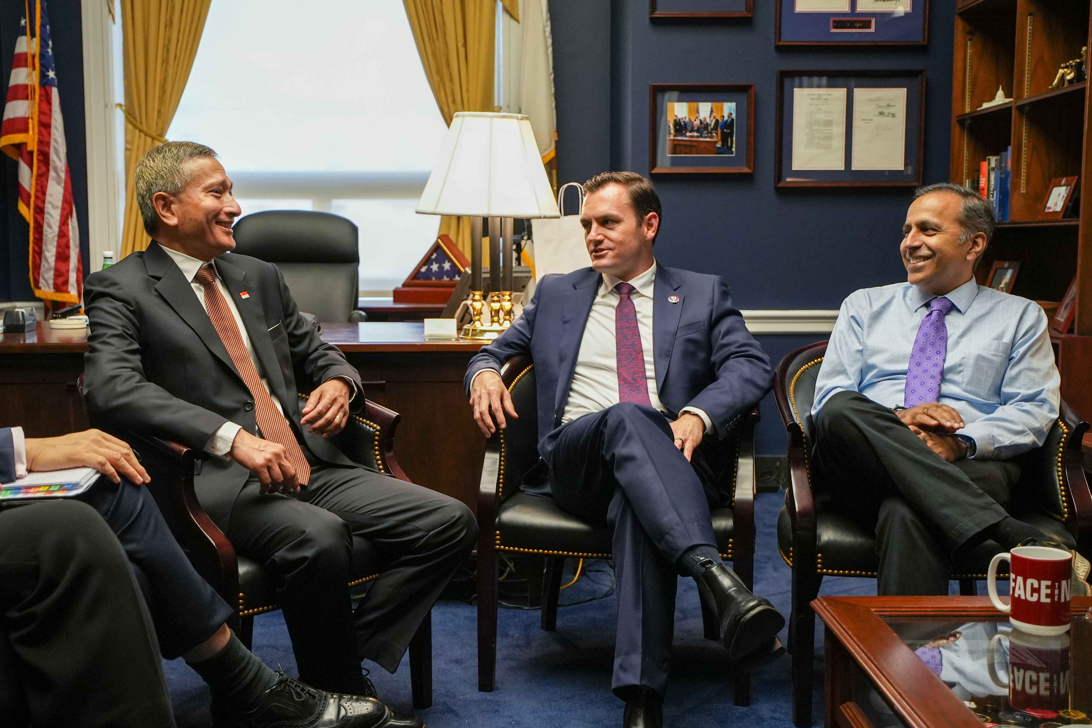 Three people in suits sit in an office, engaged in discussion. A "FACE IT" mug is on a table.